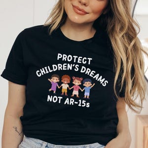 Op de afbeelding: Zwart T-shirt met de witte tekst "PROTECT CHILDREN'S DREAMS NOT AR-15s". Het ontwerp bevat vier cartoonkinderen die elkaars hand vasthouden. Het shirt heeft een ronde hals.