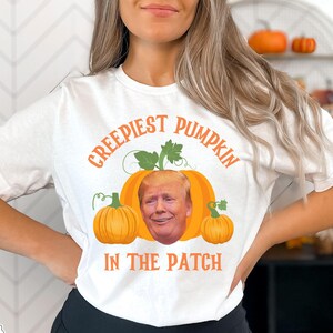May include: White t-shirt with a graphic of a pumpkin with a face that looks like Donald Trump. The text on the shirt reads "Creepiest Pumpkin in the Patch".