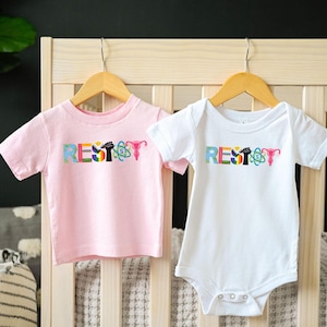 Può includere: Due body per neonati, uno rosa e uno bianco, con la parola "RESIST" scritta in lettere colorate. Ogni lettera è decorata con un simbolo diverso, tra cui un arcobaleno, un pugno, un utero e un atomo.