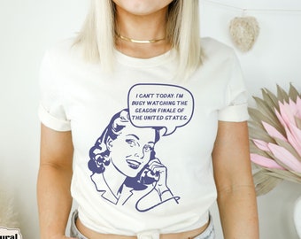 May include: White t-shirt featuring a vintage-style illustration of a woman on the phone. The text on the shirt reads "I can't today. I'm busy watching the season finale of the United States."