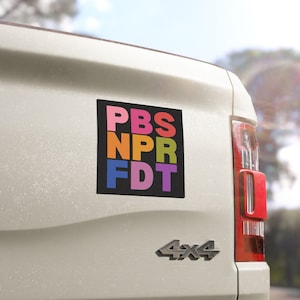 May include: A black rectangular sticker with the text "PBS NPR FDT" in colorful block letters. The sticker is affixed to the back of a white vehicle, near the taillight. The background is a sunny day.