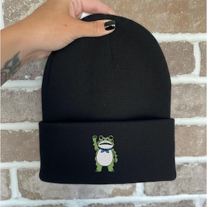 Puede incluir: Gorro negro con borde doblado. El gorro presenta un diseño de rana bordada en la parte delantera. La rana es verde y blanca, lleva una pajarita azul y saluda. El gorro está hecho de un material de punto suave.
