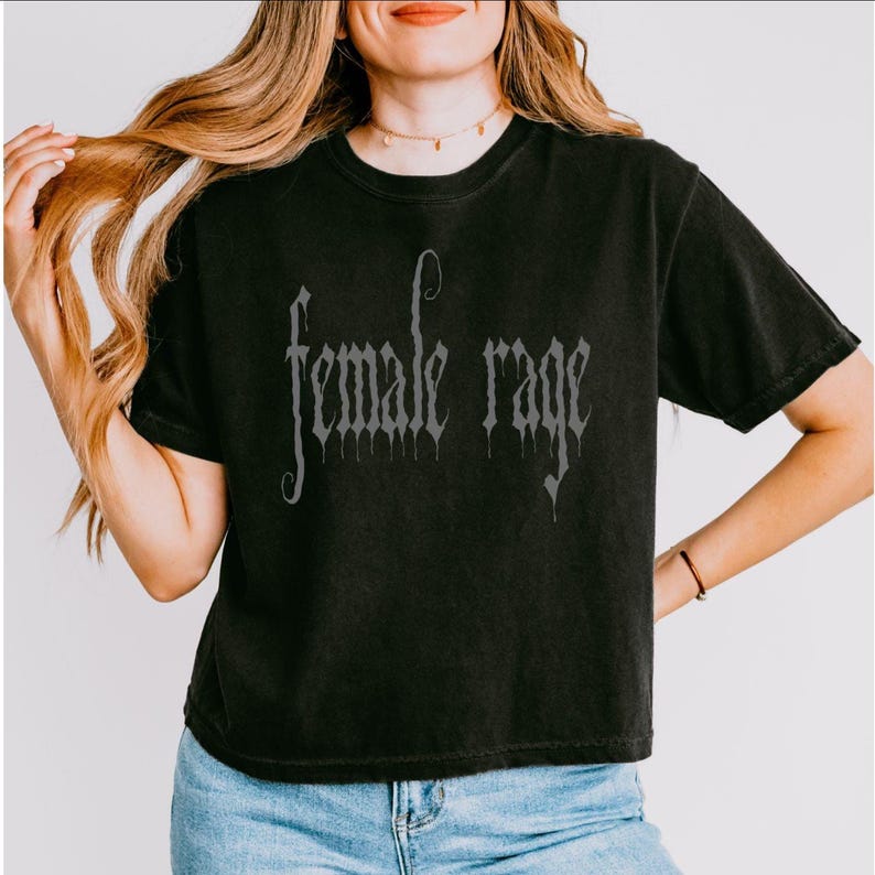 May include: Black t-shirt with the phrase "female rage" in a gothic font. The shirt is a dark color and has short sleeves. The text is in a lighter shade of gray.