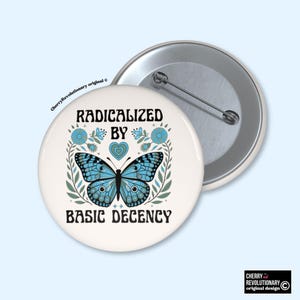 Może przedstawiać: Biała przypinka z napisem "RADICALIZED BY BASIC DECENCY" i niebieskim motylem. Motyl otoczony jest niebieskimi kwiatami i zielonymi liśćmi. Drugi przypinka widoczna w tle.