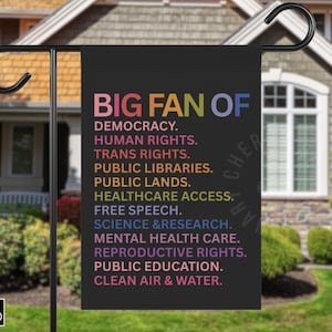 Op de afbeelding: Zwarte tuinvaandel met de kleurrijke tekst "BIG FAN OF", gevolgd door een lijst met waarden zoals democratie en mensenrechten. De vlag wordt buiten voor een huis getoond.