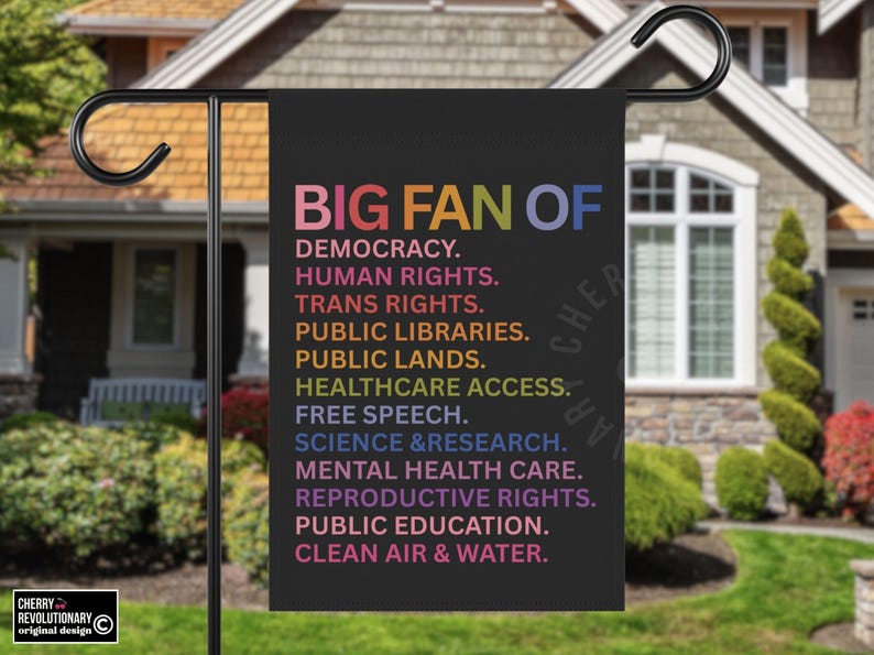 以下が含まれることがあります： 「BIG FAN OF」とカラフルな文字で書かれた黒いガーデンフラッグ。民主主義、人権、きれいな空気など、さまざまな価値観がリストされています。旗は家の前で屋外に展示されています。