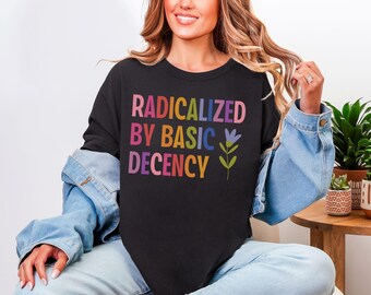 Op de afbeelding: Zwart sweatshirt met de tekst "RADICALIZED BY BASIC DECENCY" in regenboogkleuren. Een bloemenontwerp is ook inbegrepen. Onderdeel van een kledingcollectie.