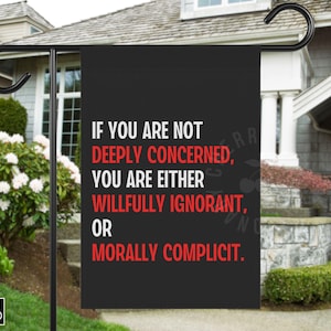 May include: Black garden flag with white and red text: "IF YOU ARE NOT DEEPLY CONCERNED, YOU ARE EITHER WILLFULLY IGNORANT, OR MORALLY COMPLICIT." The flag is hanging on a black metal stand in front of a house.