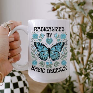 Pode incluir: Caneca de cerâmica branca com um desenho de borboleta azul e o texto "RADICALIZED BY BASIC DECENCY". A borboleta é rodeada por elementos florais e corações. A caneca tem uma pega grande.