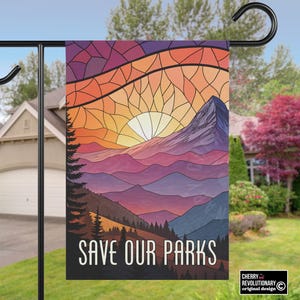 Puede incluir: Una bandera de jard&iacute;n con un dise&ntilde;o de paisaje monta&ntilde;oso y una puesta de sol en tonos naranja, amarillo y morado. El texto "SAVE OUR PARKS" se muestra en blanco. La bandera est&aacute; colgada de un poste de metal negro.