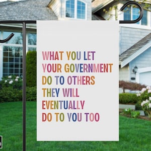 Puede incluir: Una bandera de jardín blanca con el texto "WHAT YOU LET YOUR GOVERNMENT DO TO OTHERS THEY WILL EVENTUALLY DO TO YOU TOO" en letras coloridas. La bandera se muestra al aire libre frente a una casa.