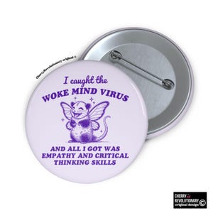 Può includere: Un bottone rotondo lilla chiaro con un'illustrazione viola di un opossum con ali di fata. Il testo recita "I caught the WOKE MIND VIRUS" e "AND ALL I GOT WAS EMPATHY AND CRITICAL THINKING SKILLS."