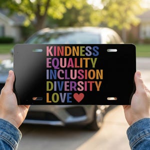 Könnte beinhalten: Schwarzes Nummernschild mit den Worten "KINDNESS, EQUALITY, INCLUSION, DIVERSITY, LOVE" in Regenbogenfarben. Ein rotes Herz steht unter dem Wort "LOVE". Das Schild wird vor einem Auto gehalten.