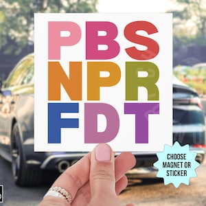 May include: A square sticker or magnet with the text "PBS NPR FDT" in colorful block letters. The background shows a car and a hand holding the item. The item is available as a magnet or sticker.