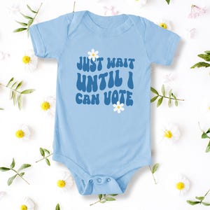 Puede incluir: Body de bebé azul claro con el texto "JUST WAIT UNTIL I CAN VOTE" en una fuente retro. El body tiene un cierre de corchetes y está decorado con margaritas. El fondo es blanco con detalles florales.