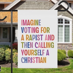 May include: A white garden flag with the text "IMAGINE VOTING FOR A RAPIST AND THEN CALLING YOURSELF A CHRISTIAN" in colorful block letters. The flag is hanging from a black metal pole in front of a house.