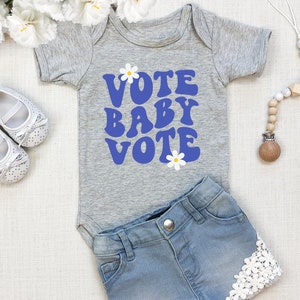 Può includere: Un body per neonato grigio con il testo "VOTE BABY VOTE" in blu con margherite bianche. Il body è abbinato a pantaloncini di jeans e un mordedor di legno.