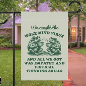 May include: A garden flag with a green and white design. The flag features two frogs holding teacups with the text "We caught the WOKE MIND VIRUS and all we got was empathy and critical thinking skills."