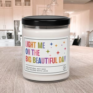 K&ouml;nnte beinhalten: Eine wei&szlig;e Kerze in einem Glas mit schwarzem Deckel. Das Etikett lautet "LIGHT ME ON THE BIG BEAUTIFUL DAY" in farbigem Text mit Sternakzenten. Die Kerze besteht aus nat&uuml;rlichem Sojawachs und ist handgegossen.