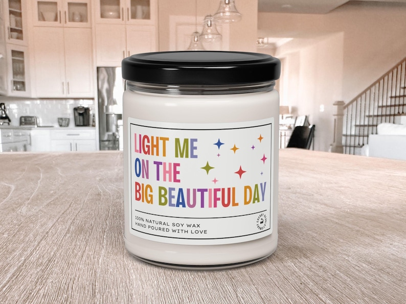 K&ouml;nnte beinhalten: Eine wei&szlig;e Kerze in einem Glas mit schwarzem Deckel. Das Etikett lautet "LIGHT ME ON THE BIG BEAUTIFUL DAY" in farbigem Text mit Sternakzenten. Die Kerze besteht aus nat&uuml;rlichem Sojawachs und ist handgegossen.