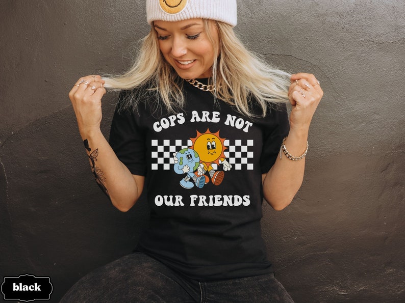 May include: A black t-shirt with white text that reads 'Cops Are Not Our Friends'. The text is in a retro font and is surrounded by a checkered pattern. A cartoon sun and earth are holding hands and walking in the checkered pattern.