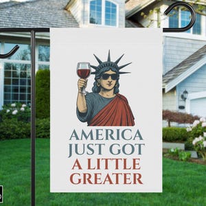 May include: A white garden flag featuring an illustration of the Statue of Liberty wearing sunglasses and holding a glass of red wine. The flag reads "AMERICA JUST GOT A LITTLE GREATER" in blue and red text.
