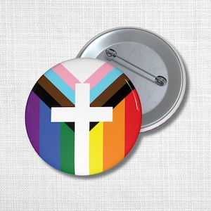 Op de afbeelding: Een ronde pin-back button met een progress pride vlagontwerp met verticale strepen in rood, oranje, geel, groen, blauw en violet, bedekt met een wit kruis en een chevronpatroon in lichtblauw, roze, wit, bruin en zwart.