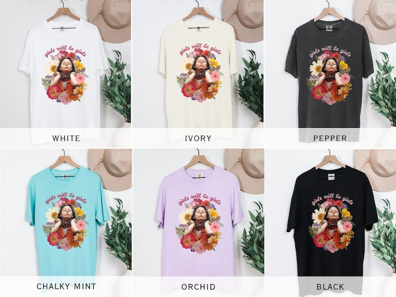 May include: A collection of t-shirts in various colors, including white, ivory, pepper, chalky mint, orchid, and black. Each shirt features a graphic of a person surrounded by flowers, with the text "girls will be girls" above the image.