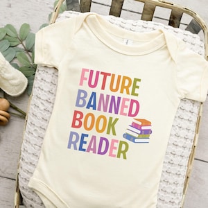 Puede incluir: Body de bebé color crema con el texto "FUTURE BANNED BOOK READER" en letras coloridas. Una pila de libros está impresa debajo del texto. El body se muestra en una cesta de mimbre con una manta blanca.