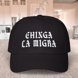 Può includere: Cappellino da baseball nero con testo ricamato bianco che recita "CHINGA LA MIGRA" in un font gotico.