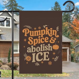 May include: A brown garden flag with the text "Pumpkin Spice & abolish ICE" in orange and beige. The flag features a pumpkin graphic and floral accents. The flag is hanging outside a house.