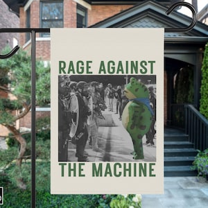 Puede incluir: Una bandera de jardín con el texto "RAGE AGAINST THE MACHINE" en verde. La bandera presenta una imagen en blanco y negro de una protesta con una mascota de rana. La bandera está colgada de un soporte de metal negro.