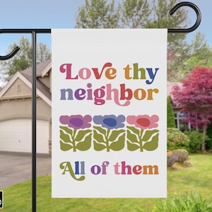May include: A white garden flag with the text "Love thy neighbor" in colorful retro font above a row of stylized flowers. The phrase "All of them" is below the flowers. The flag is hanging on a black metal pole.