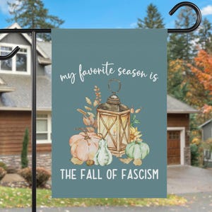 Può includere: Una bandiera da giardino turchese con la scritta "my favorite season is THE FALL OF FASCISM". La bandiera presenta un'illustrazione ad acquerello di una lanterna, zucche e fogliame autunnale.