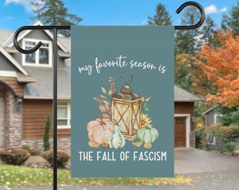 May include: A teal garden flag with the text "my favorite season is THE FALL OF FASCISM". The flag features a watercolor illustration of a lantern, pumpkins, and fall foliage.