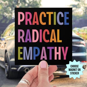 May include: A black square magnet or sticker with the text "PRACTICE RADICAL EMPATHY" in rainbow colors. The text is in all caps and the words are stacked on top of each other. The magnet or sticker is being held by a hand in front of a car.