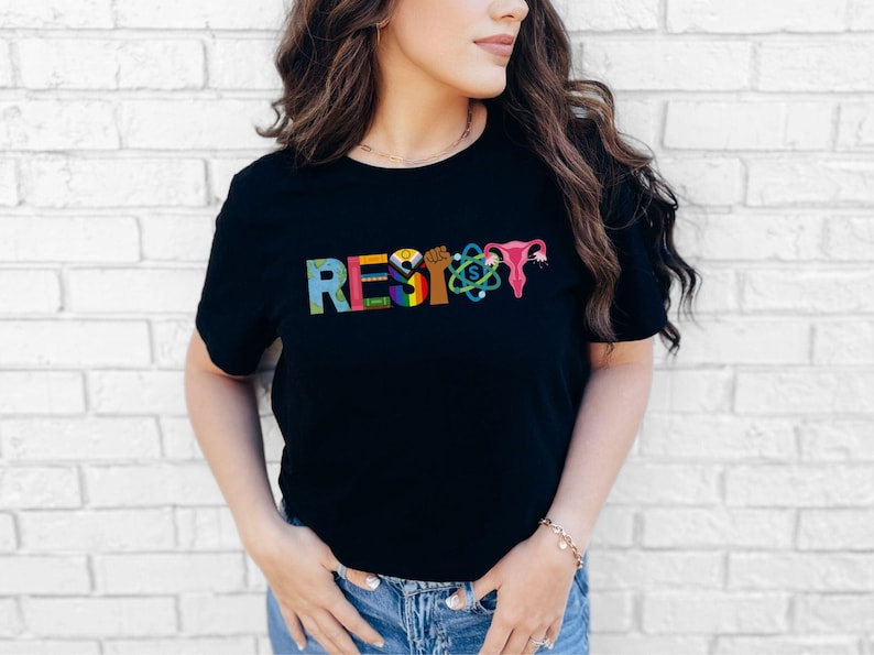 Puede incluir: Camiseta negra con la palabra "RESIST" impresa en letras de colores. Las letras est&aacute;n formadas por varios s&iacute;mbolos, incluyendo un pu&ntilde;o, un arco&iacute;ris, un &uacute;tero y un &aacute;tomo.