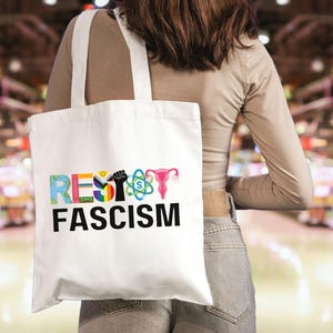 May include: White tote bag with the words "RESIST FASCISM" in colorful letters. The design includes a raised fist, a rainbow, a uterus, and an atom symbol. The bag is carried over a person's shoulder.
