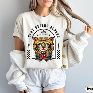 May include: White t-shirt with a tiger graphic and the text "Deny Defend Depose Eat the Rich".