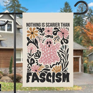 Può includere: Una bandiera da giardino con la scritta "NOTHING IS SCARIER THAN FASCISM" in nero. La bandiera presenta un fantasma rosa circondato da fiori rosa, arancioni e neri. La bandiera è appesa fuori da una casa.