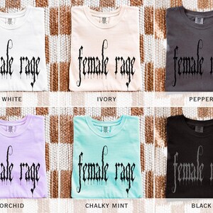 May include: Six folded t-shirts in various colors: white, ivory, pepper, orchid, chalky mint, and black. Each shirt features the text "female rage" in a gothic font. The shirts are displayed on a woven, neutral-toned surface.