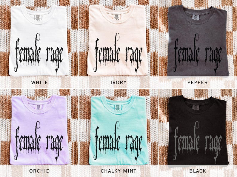 May include: Six folded t-shirts in various colors: white, ivory, pepper, orchid, chalky mint, and black. Each shirt features the text "female rage" in a gothic font. The shirts are displayed on a woven, neutral-toned surface.