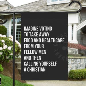 May include: Black garden flag with white text: "IMAGINE VOTING TO TAKE AWAY FOOD AND HEALTHCARE FROM YOUR FELLOW MEN AND THEN CALLING YOURSELF A CHRISTIAN." The flag is displayed on a black metal stand in front of a house.