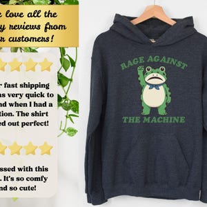 May include: Dark navy blue hoodie with a green frog graphic. The frog is raising a fist and wearing a blue bow tie. The text "RAGE AGAINST THE MACHINE" is above and below the frog in green.