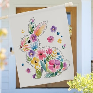 May include: A white garden flag with a watercolour bunny design filled with colourful flowers. The bunny is outlined with the words "fuck Trump". The flag is hanging on a wooden pole.