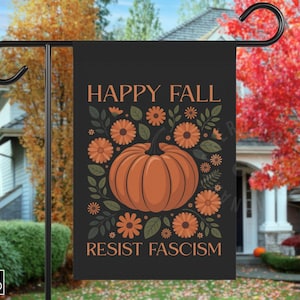 Può includere: Bandiera da giardino nera con la scritta "HAPPY FALL RESIST FASCISM" in arancione. La bandiera presenta una zucca arancione circondata da fiori arancioni e foglie verdi. La bandiera è appesa a un supporto in metallo nero.