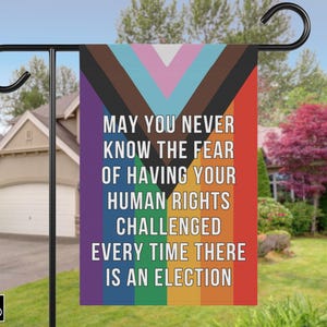 May include: A garden flag with a black pole and hook. The flag features a design with the text "MAY YOU NEVER KNOW THE FEAR OF HAVING YOUR HUMAN RIGHTS CHALLENGED EVERY TIME THERE IS AN ELECTION". The flag has a rainbow color scheme.