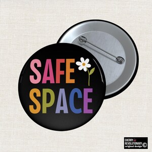 Puede incluir: Un botón negro con las palabras "SAFE SPACE" en letras coloridas y una ilustración de una margarita blanca. El botón es redondo con un alfiler en la parte posterior. El diseño incluye el texto "CHERRY REVOLUTIONARY original design".