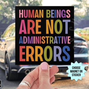 May include: A black square sticker with the text "HUMAN BEINGS ARE NOT ADMINISTRATIVE ERRORS" in rainbow colors. The sticker is being held by a hand and there is a smaller white starburst sticker below it that says "CHOOSE MAGNET OR STICKER".