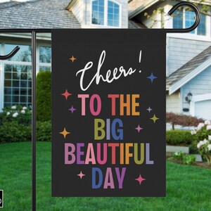 May include: A black garden flag with the words "Cheers! To The Big Beautiful Day" in white and colourful letters. The flag is hanging on a black metal pole in front of a house.
